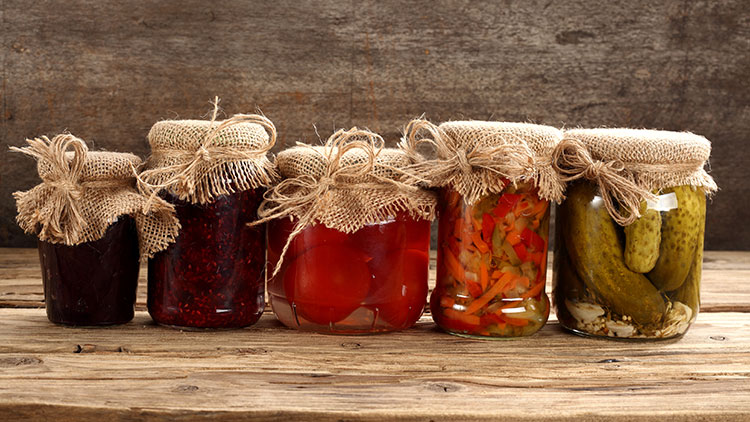 Fünf Gläser mit Stoffabdeckung und Schleifen, gefüllt mit Marmelade, Tomaten, eingelegtem Gemüse und Gurken, auf Holztisch.