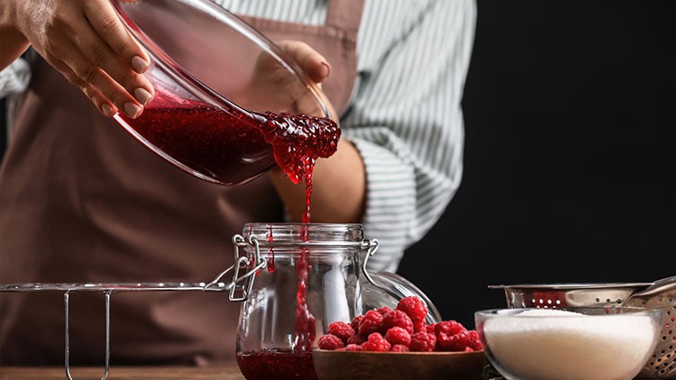 Eine Person in Schürze füllt rote Himbeermarmelade aus einer Schüssel in ein Glas, daneben frische Himbeeren und Zucker.