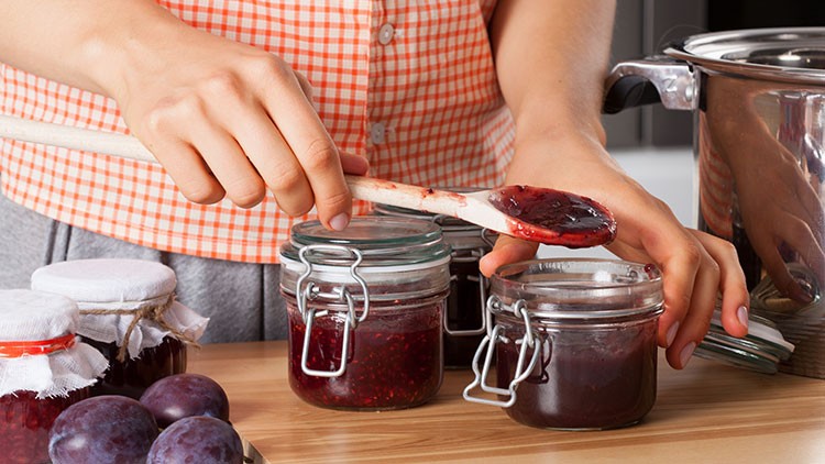 Person füllt rote Marmelade mit einem Holzlöffel in ein Bügelglas, daneben stehen Pflaumen und weitere Marmeladengläser.