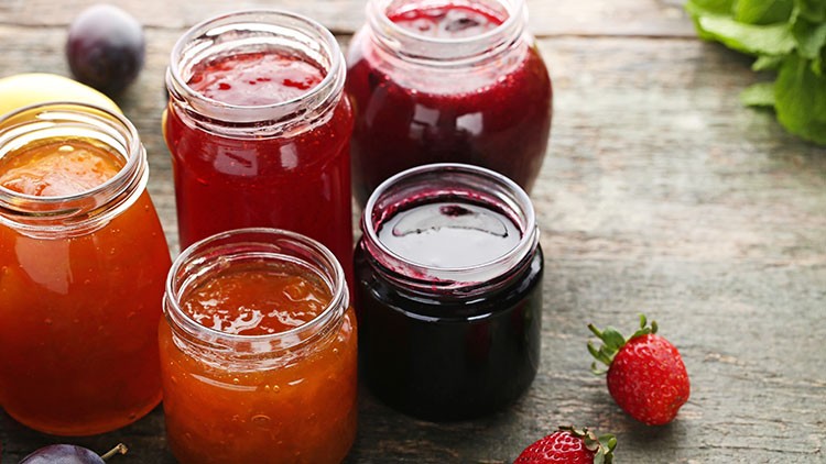 Fünf geöffnete Gläser mit roter, orangener und dunkler Marmelade stehen auf einem Holztisch, daneben Erdbeeren und Pflaumen.