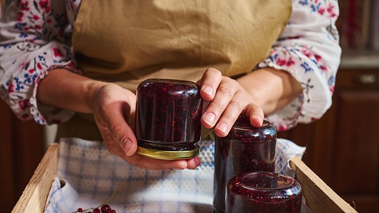 Person in Schürze stellt ein Glas mit roter Marmelade kopfüber in eine Holzkiste mit blau-weiß kariertem Tuch.