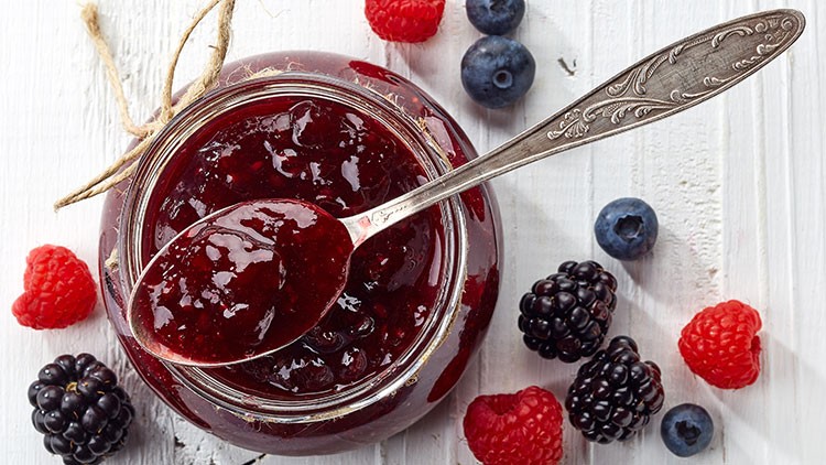 Ein Glas mit roter Beerenmarmelade, ein verzierter Löffel liegt darauf, umgeben von Himbeeren, Brombeeren und Blaubeeren.