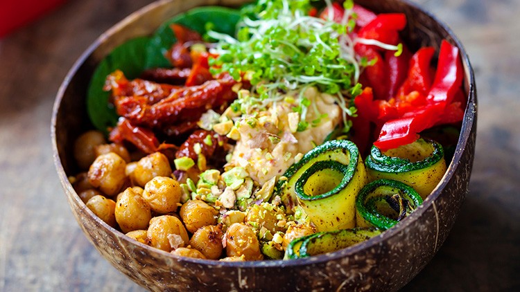 Bunte Schüssel mit Kichererbsen, Zucchinischeiben, Hummus, Sprossen, Paprika, Spinat, getrockneten Tomaten und gehackten Pistazien.