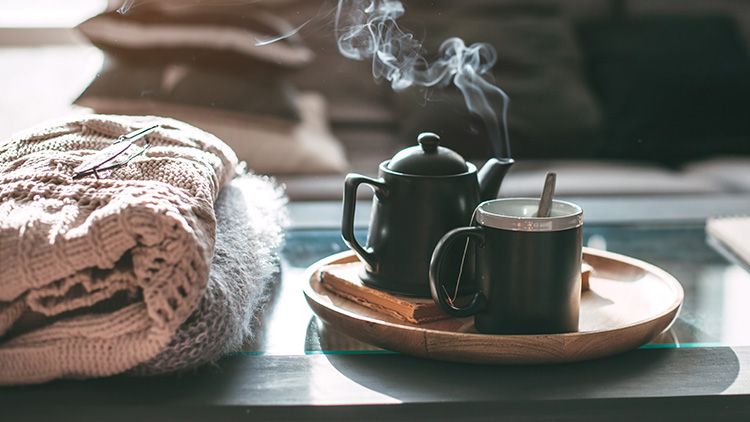 Schwarze Teekanne und Tasse auf einem runden Holztablett, daneben ein Buch und eine Decke mit Brille auf einem Glastisch.