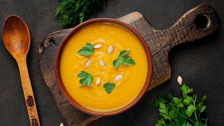 Eine Schüssel mit orangefarbener Cremesuppe, garniert mit Petersilie und Kürbiskernen, auf einem Holzbrett neben einem Holzlöffel.