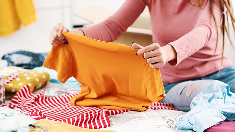 Person mit rosa Oberteil und zerrissener Jeans faltet ein orangefarbenes T-Shirt auf einem Bett, das mit Kleidung bedeckt ist.