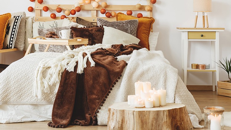 Gemütlich dekoriertes Schlafzimmer mit Kissen, Decken, Nachttisch, Kerzen auf einem Holzblock und Lichterkette am Bett.