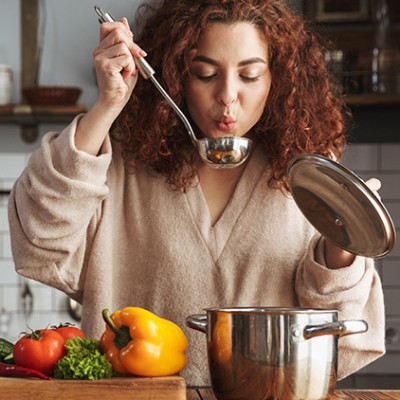 Person in beigefarbener Kleidung probiert mit einer Kelle aus einem Edelstahltopf, davor liegen Paprika, Tomaten und Brokkoli.