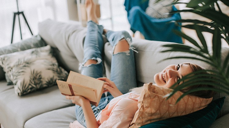 Person mit zerrissener Jeans und Buch liegt entspannt auf einem grauen Sofa, umgeben von Kissen und Pflanzen.