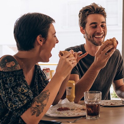 Drei Personen sitzen an einem Tisch mit Sandwiches, Cola-Gläsern und Saucenflaschen, umgeben von Tageslicht und Pflanzen.