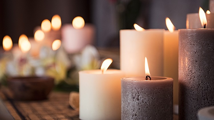 Mehrere brennende, helle und dunkle Stumpenkerzen stehen auf einem Tisch mit Blumen und unscharfem Hintergrund.