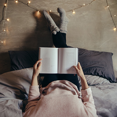 Eine Person liegt auf einem Bett, hält ein geöffnetes Buch, hat die Beine an der Wand und trägt graue Socken, umgeben von Lichterketten.