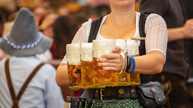 Person trägt mehrere Bierkrüge mit Schaum in beiden Händen, im Hintergrund Menschen und unscharfe Bewegungen in Festkleidung.