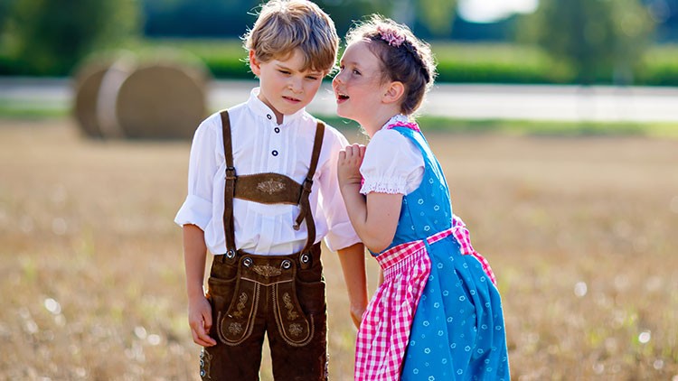 Ein Kind in Lederhose und ein Kind im blauen Dirndl stehen auf einem Feld, im Hintergrund Heuballen und Bäume.