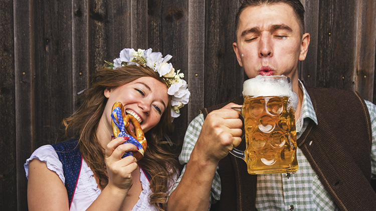 Person in Trachtenhemd hält gefüllten Bierkrug, daneben Person mit Blumenkranz und Lebkuchenherz vor Holzwand.