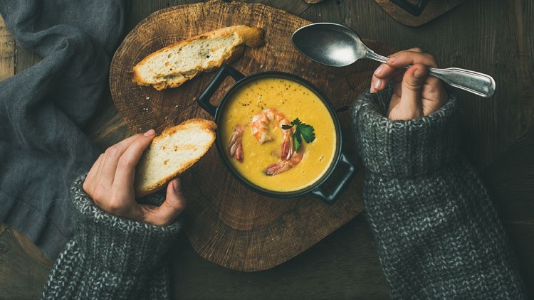 Eine Person isst Suppe mit Garnelen und Petersilie aus einem schwarzen Schälchen und hält ein Stück Brot in der anderen Hand.