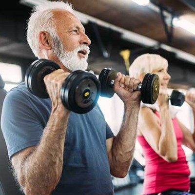 Zwei Personen heben gleichzeitig schwarze Kurzhanteln in einem Fitnessstudio mit großen Fenstern und moderner Ausstattung.