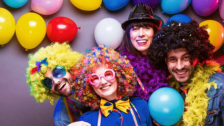 Bunte Luftballons und Personen mit farbenfrohen Perücken, Paillettenhut und gelben Federboas vor grauem Hintergrund.
