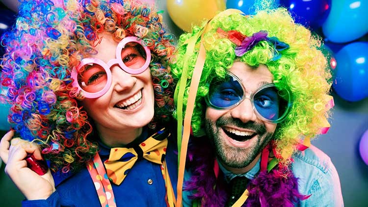 Zwei Personen mit bunten, lockigen Perücken, bunten Schleifen und Luftschlangen, im Hintergrund bunte Luftballons.