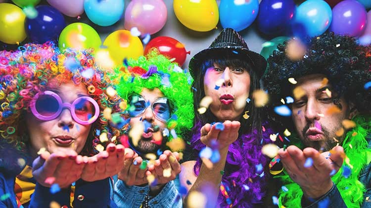 Personen in bunten Perücken und Hüten werfen Konfetti in die Luft, im Hintergrund hängen bunte Luftballons.