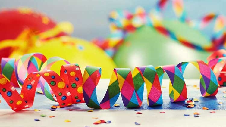 Bunte Luftschlangen, Konfetti und unscharfe Luftballons in Gelb, Rot und Grün vor blauem Hintergrund.