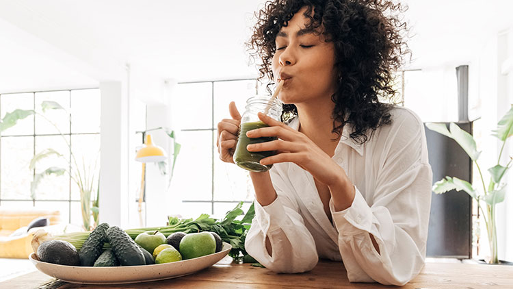 Person hält ein Glas mit grünem Smoothie in der Hand, im Hintergrund stehen Pflanzen und große Fenster lassen Licht herein.