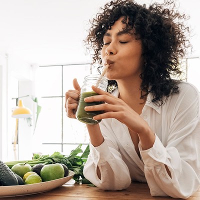 Person hält ein Glas mit grünem Smoothie in der Hand, im Hintergrund stehen Pflanzen und große Fenster lassen Licht herein.