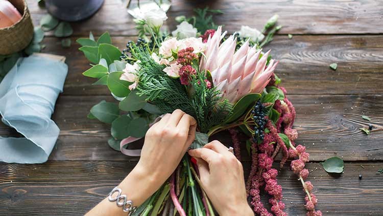 Hände binden einen Blumenstrauß mit rosa Protea, Eukalyptus und weiteren Blumen, auf einem Holztisch mit Deko-Elementen.
