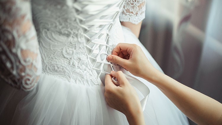Hände schnüren ein weißes Kleid mit Spitzenoberteil und Schnürung am Rücken zu.