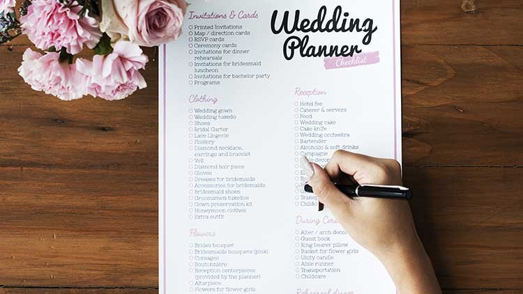 Eine Hand hält einen schwarzen Stift und hakt Punkte auf einer Wedding-Planner-Checkliste ab, daneben rosa Blumen auf Holz.