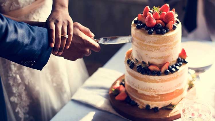 Zwei Hände halten gemeinsam ein Messer und schneiden eine zweistöckige Torte mit Erdbeeren und Heidelbeeren an.
