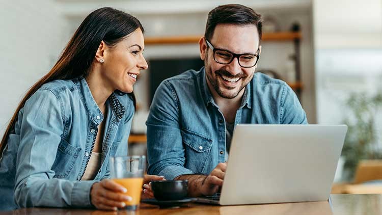 Zwei Personen in Jeanskleidung sitzen an einem Tisch mit Laptop, Orangensaftglas und einer schwarzen Tasse.