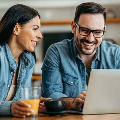 Zwei Personen in Jeanskleidung sitzen an einem Tisch mit Laptop, Orangensaftglas und einer schwarzen Tasse.