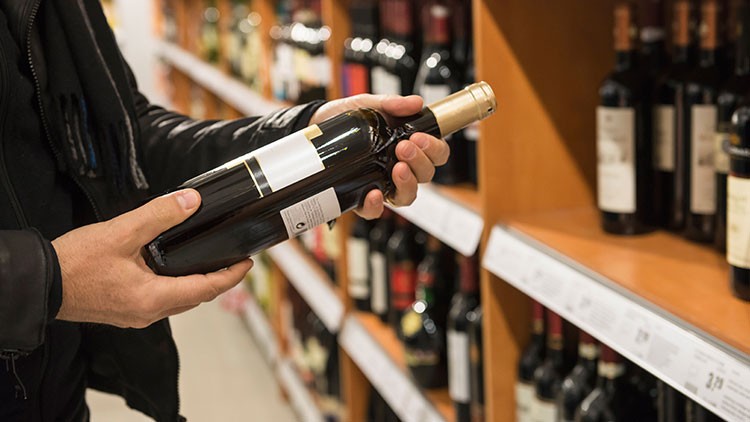 Person hält eine Weinflasche mit goldfarbenem Verschluss vor einem Regal mit weiteren Flaschen in einem Geschäft.