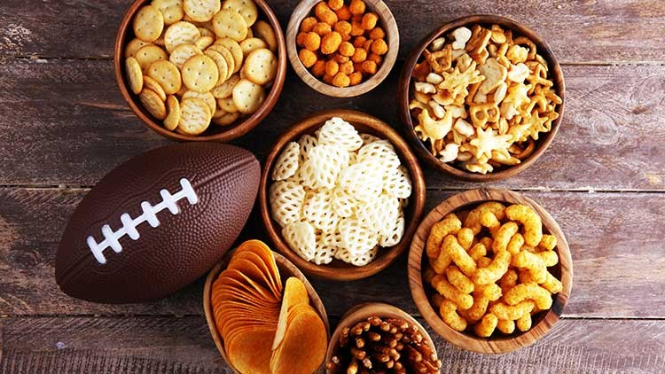 Ein brauner Football liegt neben Schalen mit Crackern, Chips, Brezeln, Käseflips und anderen Snacks auf einem Holztisch.