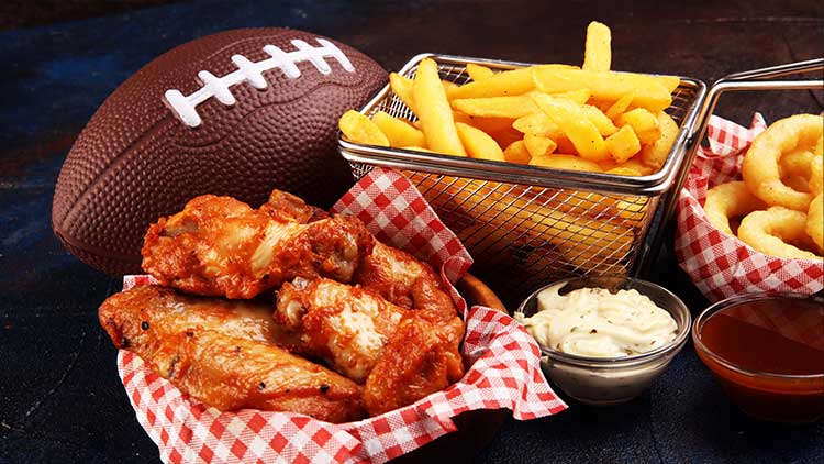 Ein Football liegt neben knusprigen Pommes, Zwiebelringen und würzigen Chicken Wings mit Dip in roten Körbchen.