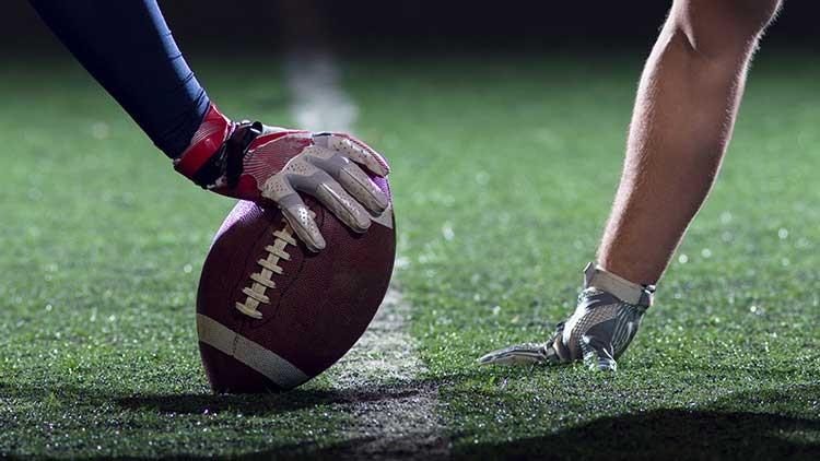 Eine behandschuhte Hand hält einen Football auf einem beleuchteten Spielfeld, daneben stützt sich eine andere Hand ab.