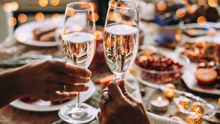 Zwei Hände stoßen mit Gläsern Sekt an, im Hintergrund ein festlich gedeckter Tisch und unscharfe Lichter.