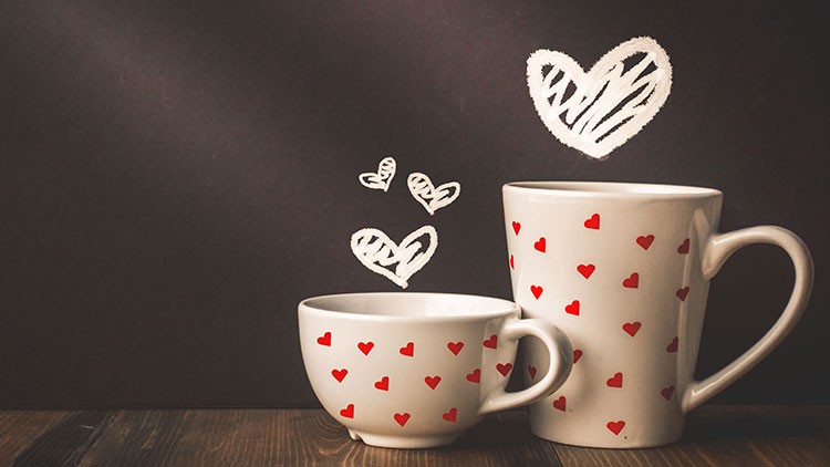 Eine weiße Tasse und eine weiße Schale mit roten Herzen stehen auf Holz vor einer schwarzen Wand mit gezeichneten weißen Herzen.
