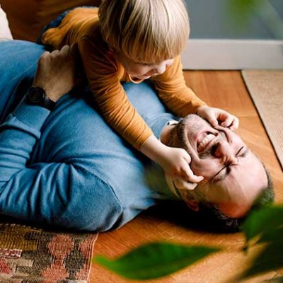 Eine Person liegt lachend auf einem Holzboden, während ein Kind spielerisch dessen Gesicht berührt, Teppich und Pflanze im Vordergrund.