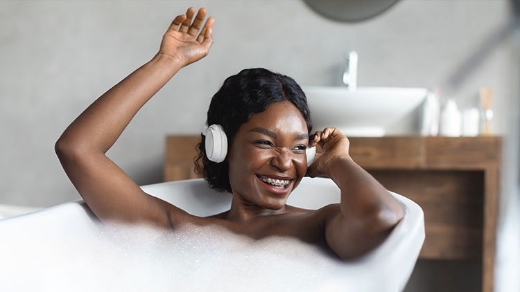 Person entspannt in einer Badewanne mit Schaum, trägt weiße Kopfhörer, im Hintergrund ein Waschbecken mit Accessoires.