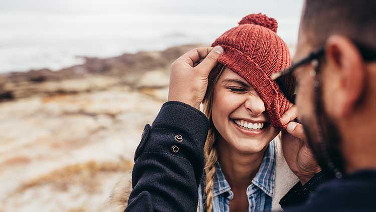 Person setzt einer anderen Person eine rote Strickmütze auf, im Hintergrund ist eine felsige Landschaft zu sehen.