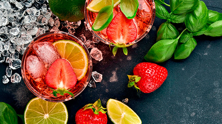 Zwei Gläser mit roten Getränken, Eiswürfeln, Erdbeeren, Limettenscheiben und Basilikumblättern, daneben frische Erdbeeren und Eiswürfel.