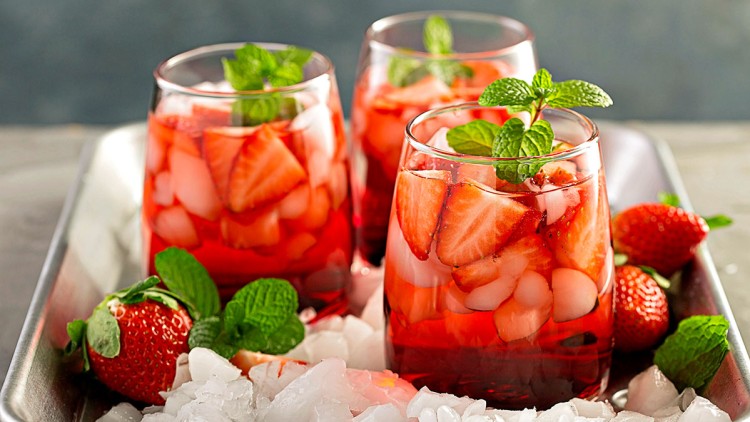Drei Gläser mit Erdbeerstückchen, Eiswürfeln, roter Flüssigkeit und frischer Minze auf Metalltablett mit weiteren Erdbeeren.