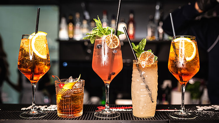 Fünf unterschiedlich dekorierte Cocktails in Gläsern mit Eiswürfeln, Zitrusscheiben, Minze und Strohhalmen auf einer Bar.