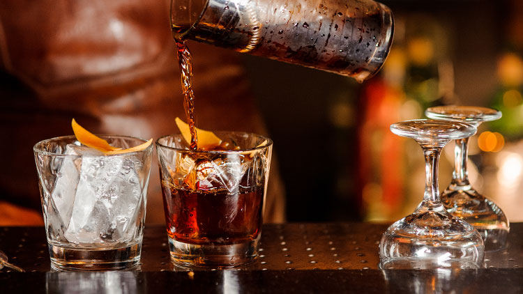 Ein Glas mit Eis und Zeste steht neben einem Glas, in das braune Flüssigkeit gegossen wird, daneben umgedrehte Weingläser.