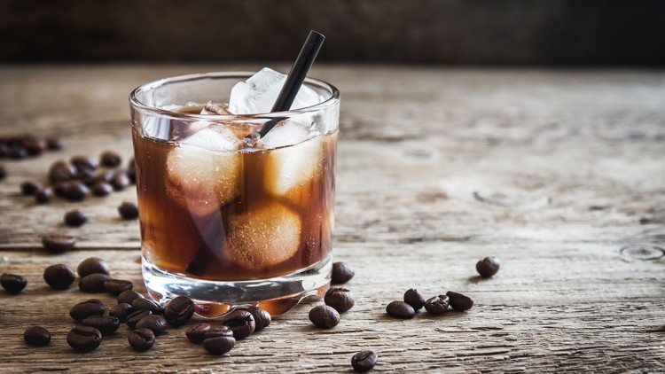 Ein Glas mit dunklem Getränk, Eiswürfeln und schwarzem Strohhalm steht auf Holztisch, umgeben von verstreuten Kaffeebohnen.