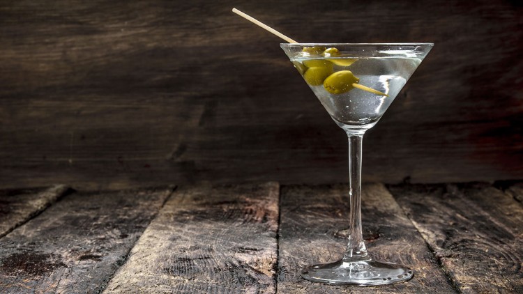 Ein gefülltes Martini-Glas mit drei grünen Oliven auf einem Holzspieß steht auf einer rustikalen Holzoberfläche.