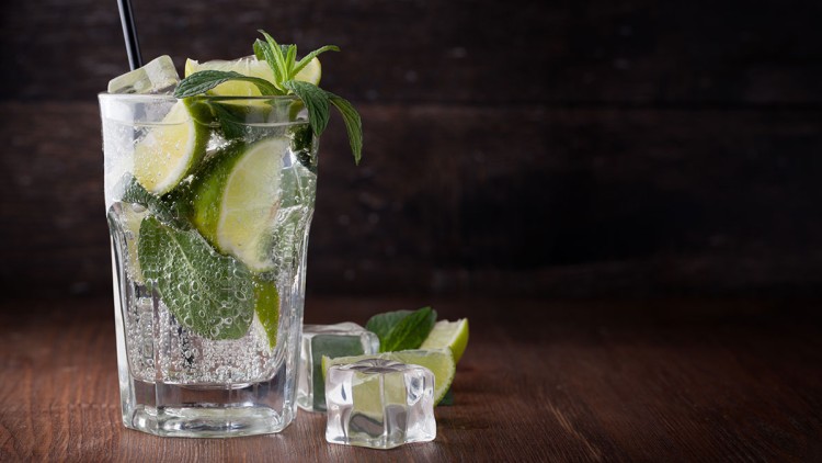 Ein Glas mit sprudelndem Wasser, Limettenscheiben, Minzblättern, Eiswürfeln und einem schwarzen Trinkhalm auf Holzuntergrund.
