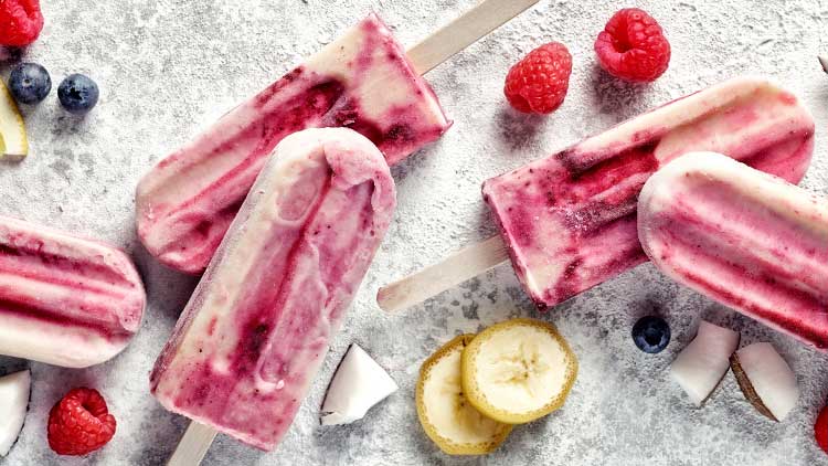 Gefrorene Fruchteiscreme am Stiel in Rosa- und Weißtönen mit Himbeeren, Blaubeeren, Zitrone und Bananenscheiben dekoriert.